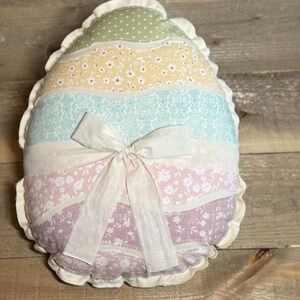 May include: A decorative egg-shaped pillow with a ruffled edge. The pillow features horizontal stripes in pastel colors: green with polka dots, yellow with small flowers, light blue with a floral pattern, white, pink with flowers, and lavender. A white bow is tied in the center.