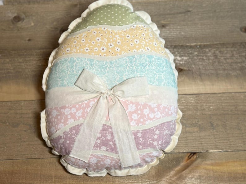 May include: A decorative egg-shaped pillow with a ruffled edge. The pillow features horizontal stripes in pastel colors: green with polka dots, yellow with small flowers, light blue with a floral pattern, white, pink with flowers, and lavender. A white bow is tied in the center.