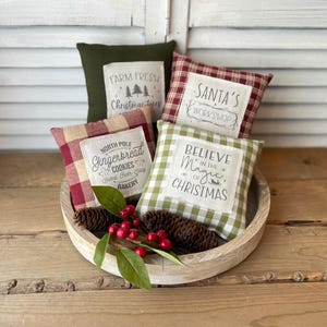 May include: Four decorative throw pillows with a Christmas theme. The pillows are in a wooden bowl. The pillows are red and green plaid with white text. The text on the pillows reads "Farm Fresh Christmas Trees", "Santa's Workshop", "North Pole Gingerbread Cookies Baked Fresh Daily Bakery", and "Believe in the Magic of Christmas".
