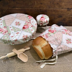 May include: A collection of kitchen textiles with a floral patchwork design. Includes a bread bag, bowl cover, jar cover, wooden rolling pin, and scoop. The textiles feature a white background with pink and red roses and green accents.
