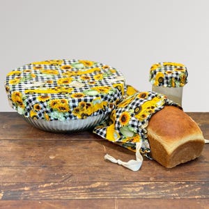 May include: A set of reusable food covers with a sunflower and black and white checkered pattern. The set includes a bowl cover, a bread bag, and a jar cover. The bread bag holds a loaf of bread.