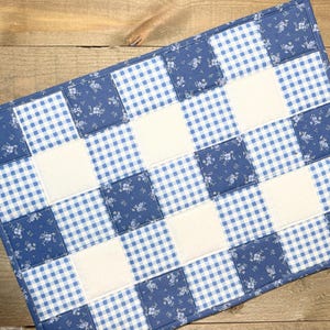 May include: A rectangular quilted placemat with a patchwork design. The pattern features alternating squares of blue gingham, white squares, and blue floral fabric. The placemat is approximately 18 inches long and 12 inches wide.