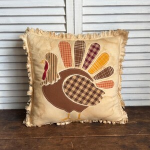 May include: Square decorative pillow with a turkey applique. The turkey is brown with a plaid body and colorful plaid tail feathers. The pillow has a cream-colored background and a fringed edge.