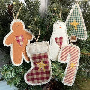 May include: A collection of Christmas ornaments in various shapes, including a gingerbread man, snowman, stocking, candy cane, and Christmas tree. Each ornament features a frayed edge and a heart or star design. The ornaments are hanging on a Christmas tree.