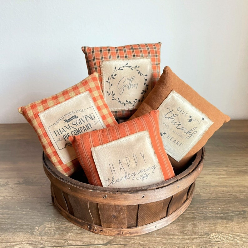 May include: Four decorative throw pillows with Thanksgiving-themed designs in a brown wicker basket. The pillows are made of a soft, textured fabric and feature a variety of colors, including orange, brown, and cream. The designs include "Gather", "Baked Fresh Daily", "Happy Thanksgiving", and "Give Thanks".