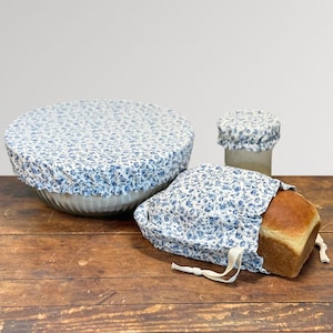 May include: A set of reusable food covers and a bread bag. The covers and bag are made of fabric with a blue floral pattern. A loaf of bread is in the bag. The items are on a wooden surface.
