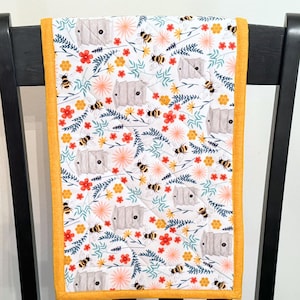 May include: A white quilted table runner with a repeating pattern of bees, beehives, and flowers in red, yellow, and peach. The runner has a yellow border and is draped over a black chair.