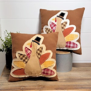 May include: Two brown decorative pillows with appliqued turkey designs. Each turkey has a tan body, a black pilgrim hat, and a colorful tail with plaid and solid fabric accents. The pillows are on a wooden surface.