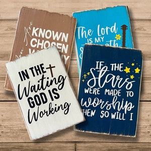 May include: Four wooden signs with inspirational quotes. The signs are painted in various colors, including blue, brown, white, and dark blue. The quotes are: "You Are Known Chosen Worthy", "The Lord Is My Strength", "In The Waiting God Is Working", and "If The Stars Were Made To Worship Then So Will I".