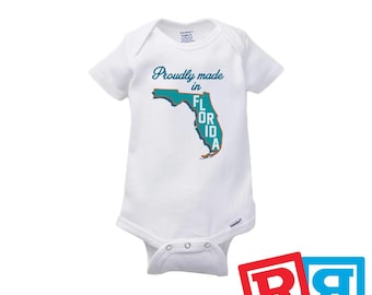 May include: A white short-sleeved baby bodysuit featuring a teal graphic of the state of Florida with the word FLORIDA written vertically in white letters inside the shape. Above the state, the text Proudly made in is printed in teal script.