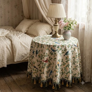 May include: A round table covered with a cream-colored tablecloth featuring a pattern of colorful birds and green branches. The tablecloth has a dark green border with hanging tassels. A brass lamp and a vase of flowers sit on top.