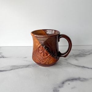 May include: A brown and tan ceramic mug with a raised floral design. The mug has a slightly flared base and a rounded handle.