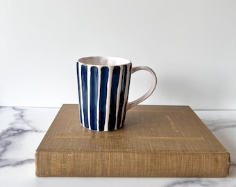 May include: A ceramic mug with a white interior and handle, featuring vertical navy blue stripes. The mug is placed on a vintage book with gold lettering. The background is a plain white wall.