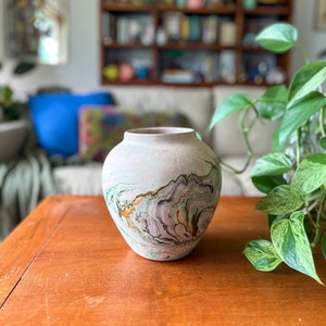 May include: A white ceramic vase with a marbled design in shades of green, brown, and black. The vase is sitting on a wooden table.