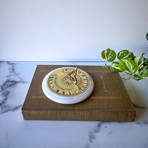 May include: A brass sundial with Roman numerals and a compass rose design, set on a white marble base. The sundial is sitting on top of a brown book.