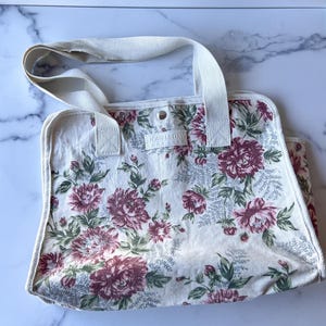 May include: Cream-colored tote bag with a floral pattern in shades of burgundy, pink, and green. The bag has a white handle and trim, and the text "DOUBLE DAY COT CLUB" is visible. The bag is rectangular with a snap closure.