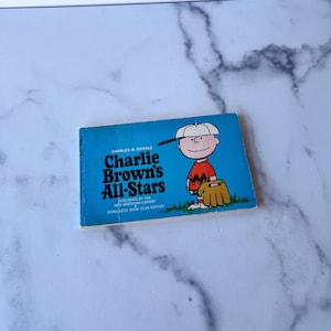 May include: A vintage copy of "Charlie Brown's All-Stars" by Charles M. Schulz. The book features a blue cover with the title and a cartoon illustration of Charlie Brown in a baseball uniform. Published by the New American Library.