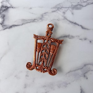 May include: A copper-colored metal hot pad with a decorative design of two stylized brooms and leaves.