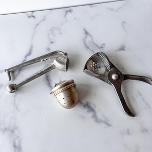 May include: An assortment of vintage silver tea infusers and a garlic press. The tea infuser is a small, perforated metal sphere with a lid. The garlic press and tea infuser are made of silver metal.