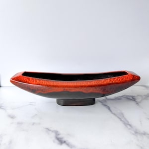 May include: A long, boat-shaped ceramic vase with a glossy, two-tone finish. The top rim is a vibrant red, transitioning to a deep brown base. The vase sits on a rectangular, dark brown base.