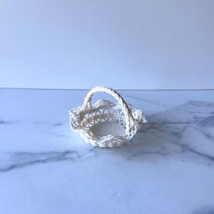 May include: A small, white woven basket with a handle. The basket has an open weave design and a round base. The handle is made of twisted rope. The basket is sitting on a marble surface.