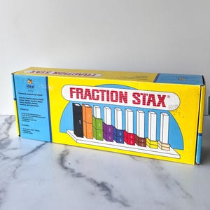 May include: A yellow box of "Fraction Stax" educational toy. The box features the product name in red letters and a diagram of colorful fraction pieces. The box is on a white surface.