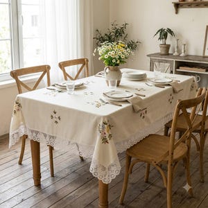 Könnte beinhalten: Eine cremefarbene, rechteckige Tischdecke mit zarter Blumenstickerei und einem breiten weißen Spitzenrand. Die Tischdecke liegt auf einem Holztisch, der mit weißen Tellern, klaren Gläsern und einer Vase mit Margeriten gedeckt ist.