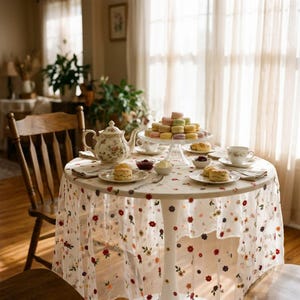 May include: A round table covered with a white sheer tablecloth featuring small embroidered red, orange, and purple flowers. On the table sits a floral teapot, a tiered stand with colourful macarons, scones, and teacups.