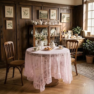 May include: A round table covered with a pale pink lace tablecloth featuring an intricate floral pattern. The table is set with a white ceramic teapot, a matching teacup and saucer, and a small plate holding two pastries, centred in a room.