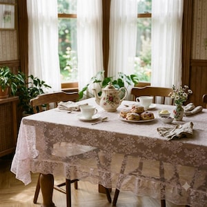 May include: A dining table covered with a light pink lace tablecloth featuring a floral pattern. On the table are a porcelain teapot with floral motifs, white teacups, a plate of scones, a small vase of wildflowers, and folded napkins.