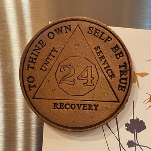Könnte beinhalten: Ein brauner Holz-Chip für 24 Stunden Abstinenz mit dem Text "To Thine Own Self Be True" und "Unity, Service, Recovery" um ein Dreieck herum. Der Chip hat die Nummer 24 in der Mitte.