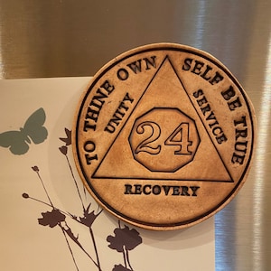 Op de afbeelding: Een 24-uurs sobriëteit chip met een driehoekig ontwerp. De driehoek heeft de woorden "To Thine Own", "Unity", "Recovery", "Self Be True" en "Service" erop gegraveerd. De chip is gemaakt van bruin materiaal en heeft een gouden afwerking.