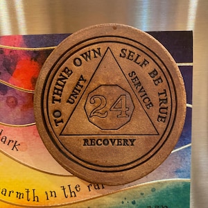 Op de afbeelding: Een bruine leren munt met de woorden "To Thine Own Self Be True" en "Unity, Service, Recovery" rond een driehoek met het nummer 24 erin.