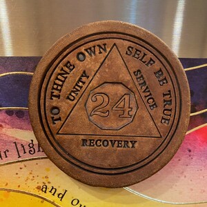Op de afbeelding: Een bruine leren onderzetter met een driehoekig ontwerp en de tekst "To Thine Own Self Be True" en "Unity, Service, Recovery". Het nummer 24 staat in het midden van de driehoek.