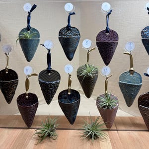 Op de afbeelding: Een display van 14 keramische hangpotten in verschillende kleuren en patronen. Elke pot heeft de vorm van een kegel en een kleine opening aan de bovenkant om op te hangen. De potten zijn in rijen op een houten plank gerangschikt.