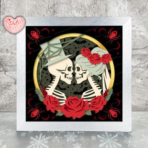 May include: Framed artwork depicting a skeleton couple kissing, encircled by red roses and spiders. One skeleton wears a top hat, the other a floral headpiece. The background is black, with a gold-colored circle.