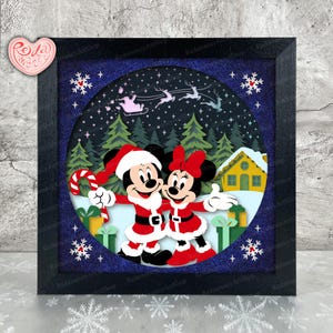 May include: A framed Christmas-themed artwork featuring Mickey and Minnie Mouse in Santa outfits, holding hands. The background shows a snowy forest, a house, and Santa's sleigh. The artwork is in a black frame.