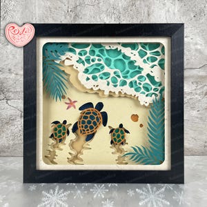 Puede incluir: Una obra de arte en papel enmarcada que representa una escena de playa con tres tortugas marinas nadando hacia el océano. La obra presenta capas de papel en tonos de azul, beige y verde, con un marco negro. También se ven una estrella de mar y conchas.