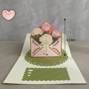 May include: A pink pop-up greeting card with an envelope design, a bow, and heart details. The envelope contains paper flowers in pink and cream, with lavender accents. The card measures 11.51 cm in height.