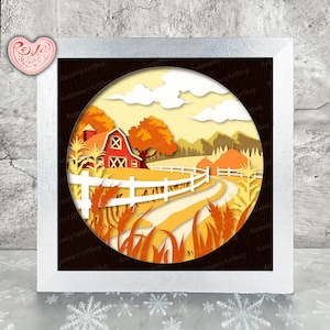 May include: A layered paper art piece depicting a fall farm scene. A red barn, white fence, and winding road are set against a yellow and orange landscape. The artwork is framed in a white, square frame.
