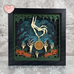 May include: A layered paper shadow box art piece in a black frame featuring a skeleton hand holding a gold pocket watch. The scene includes orange jack-o'-lanterns, skeletal hands rising from the ground, and dark trees against a blue background.