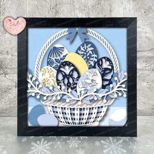 Puede incluir: Una obra de arte en papel enmarcada que presenta una cesta llena de huevos de Pascua decorados. Los huevos son en tonos de azul, blanco y amarillo, con intrincados diseños florales y geométricos. La cesta es blanca, sobre un fondo azul claro.