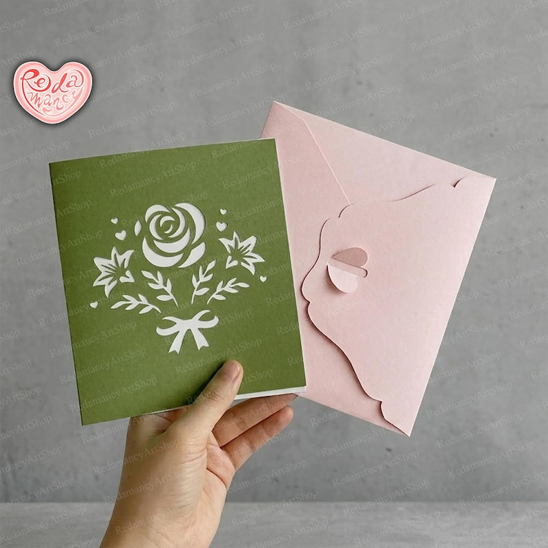 May include: A green pop-up card with a white floral design, including a rose, leaves, and a bow, is held next to a pink envelope. The card is square-shaped and features intricate cut-out details.