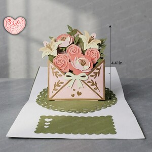 May include: A pop-up greeting card with a pink envelope design, a bow, and a floral arrangement. The card, measuring 11.2 cm tall, features pink roses, white lilies, and green leaves. The card is on a green and white base.