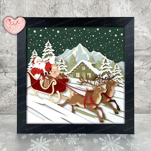 May include: A framed paper art piece depicting Santa Claus in a red sleigh pulled by two reindeer, set against a snowy winter landscape with a cabin and pine trees. The artwork is in a black frame.