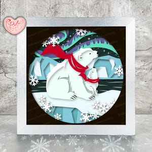 Könnte beinhalten: Gerahmtes Papierkunstwerk mit einem Eisbären und einem Jungtier mit roten Schals, vor einer Winterlandschaft mit Schneeflocken und Nordlichtern. Das Kunstwerk befindet sich in einem weißen Rahmen.