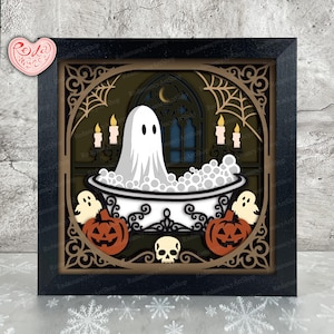 May include: A layered paper shadow box art piece in a black frame featuring a white ghost sitting in a bathtub filled with bubbles. The scene includes orange jack-o'-lanterns, a skull, lit candles, and a crescent moon against a dark background.