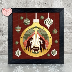 Può includere: Un design di ornamento natalizio incorniciato con una scena della natività all'interno di una forma di ornamento dorata. L'arte della carta a strati include sagome di Maria, Giuseppe e Gesù bambino, con ornamenti sospesi rossi, verdi e bianchi. La cornice è nera.