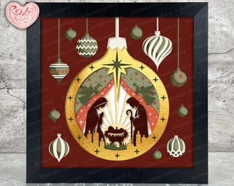 Puede incluir: Un diseño de adorno navideño enmarcado con una escena de la Natividad dentro de una forma de adorno dorada. El arte en papel en capas incluye siluetas de María, José y el niño Jesús, con adornos colgantes en rojo, verde y blanco. El marco es negro.