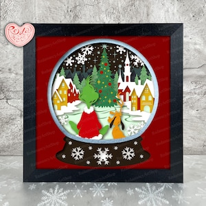 May include: A framed, layered paper art piece featuring a Christmas scene inside a snow globe. The design includes a green character in a red coat, a yellow dog, a Christmas tree, and houses. The frame is black, and the background is red.